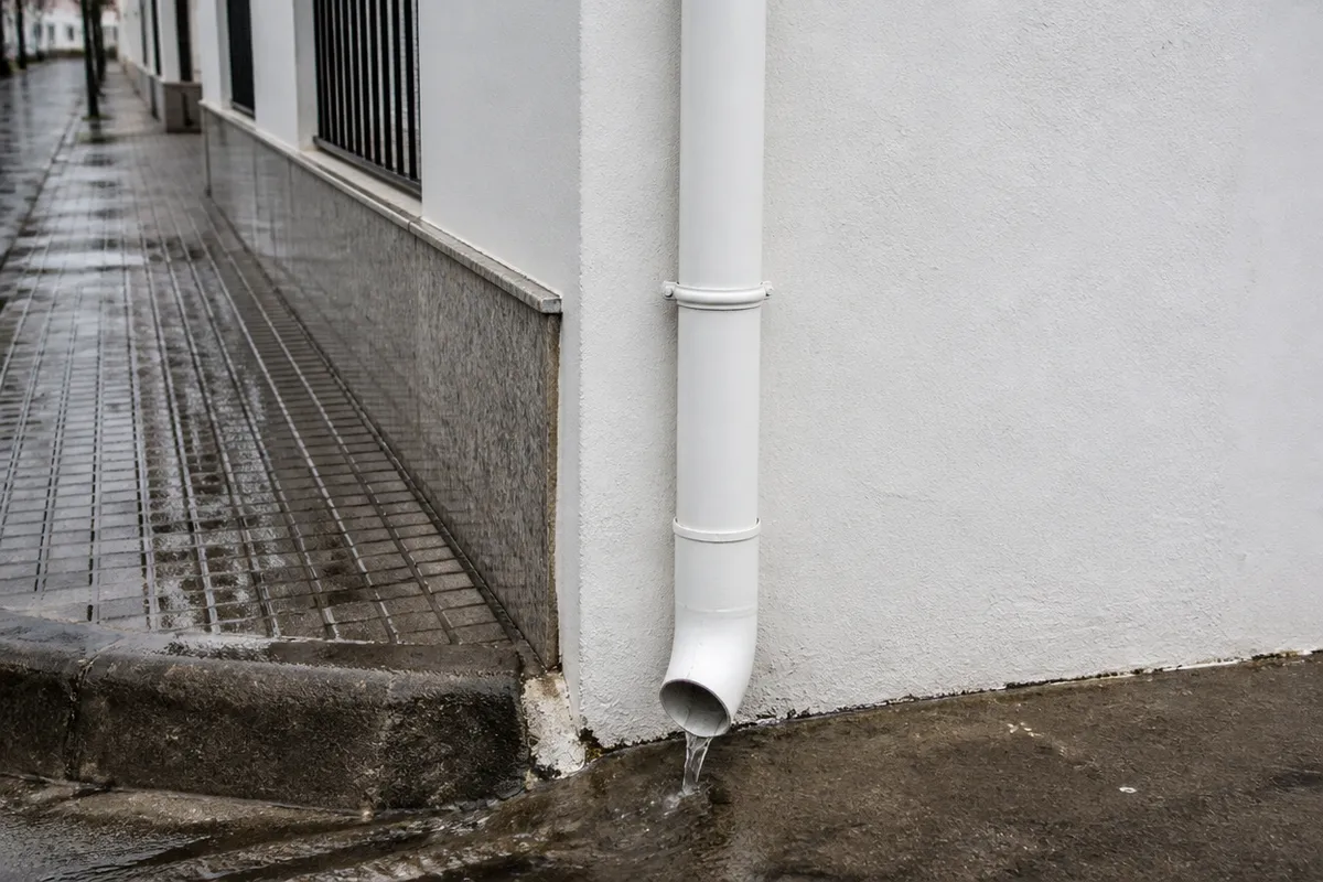 Canalón y bajante limpios en la esquina de una fachada encalada, con agua de lluvia bajando por el bajante